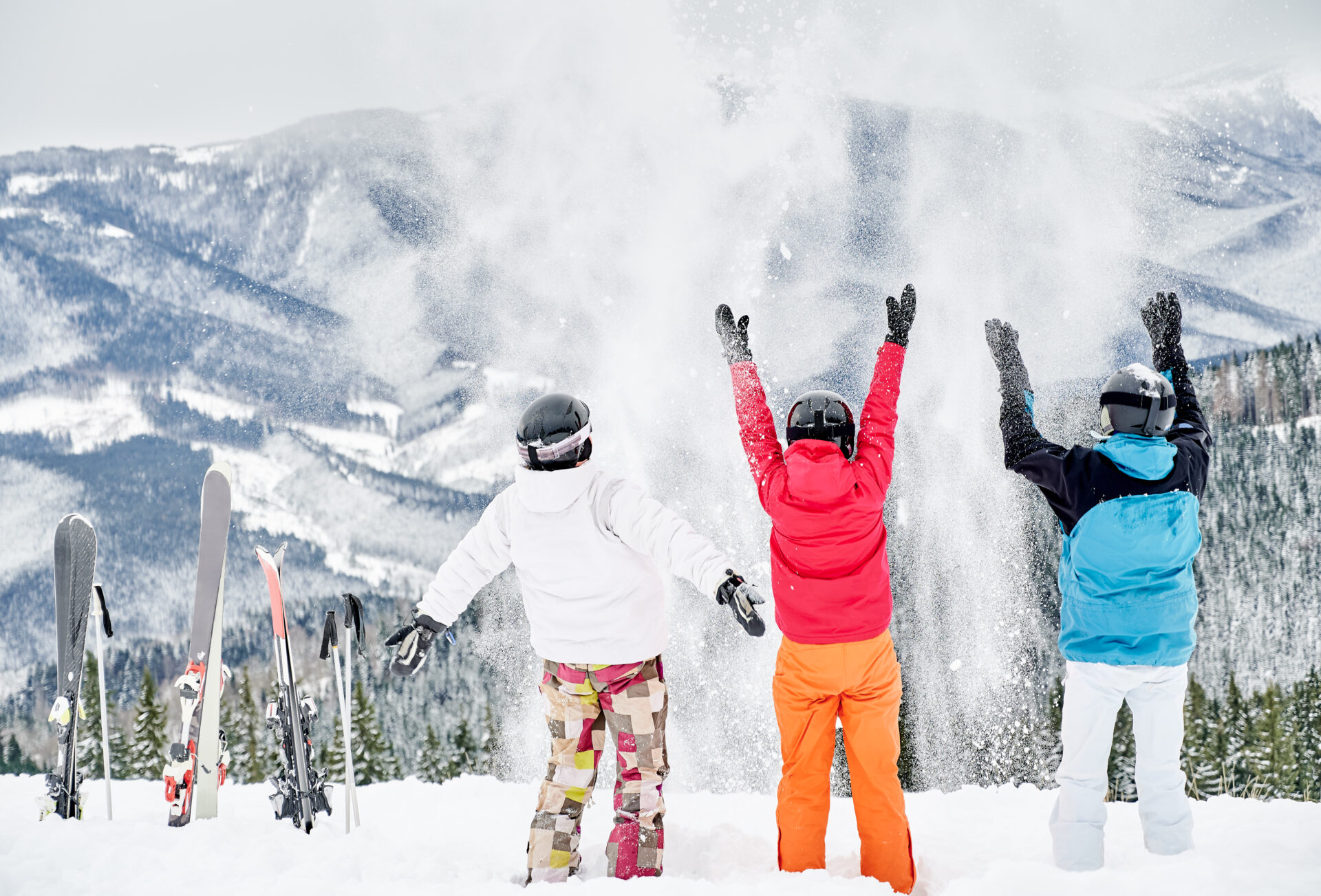 Back view of friends skiers in ski suits and helmets throwing fresh powder snow high in the air, having fun on snowy hill with beautiful mountains on background. Concept of ski resort and friendship.